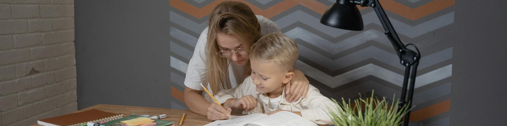 A supportive parent encouraging a primary school child who is concentrating on an open notebook during homework time, illustrating independent learning.